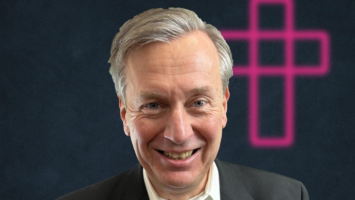 Ein lächelnder Mann im Anzug mit einer pinkfarbenen Kreuzgrafik hinter ihm.