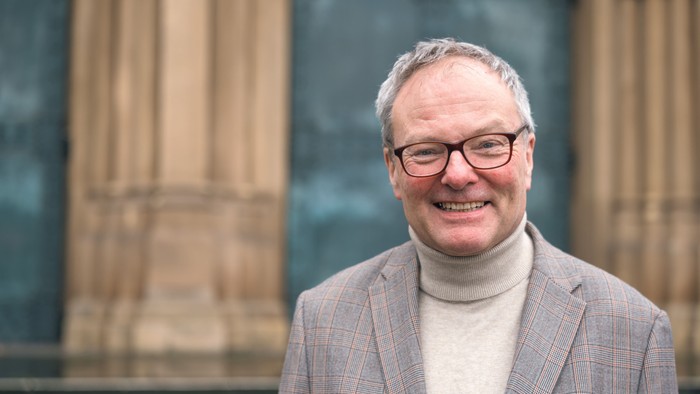 Ein lächelnder Mann mit Brille, der eine Tweedjacke und einen Rollkragenpullover trägt, vor einer strukturierten Steinmauer.