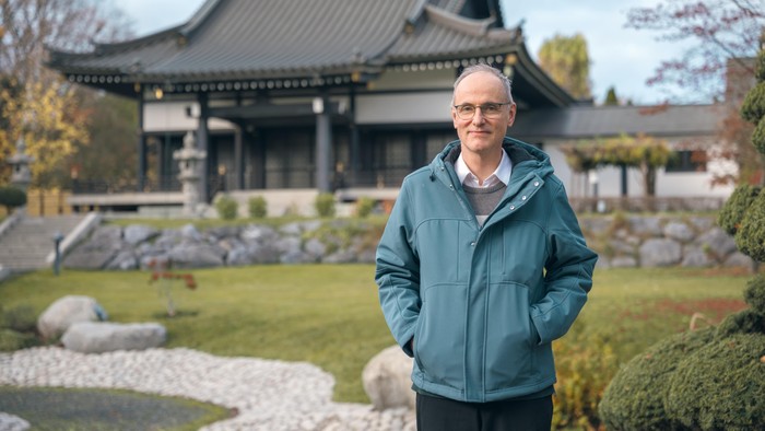 Ein Mann steht in einem ruhigen Garten mit traditioneller asiatischer Architektur im Hintergrund und Felslandschaftsgestaltung.