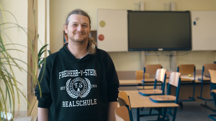 Eine Person in einem schwarzen „Freiherr-v-Stein-Realschule“-T-Shirt steht in einem Klassenzimmer mit Tischen und einer Projektionsleinwand.