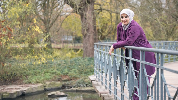 Eine Person in einem lila Hidschab und Mantel steht lächelnd auf einer Metallbrücke neben einem ruhigen Bach.