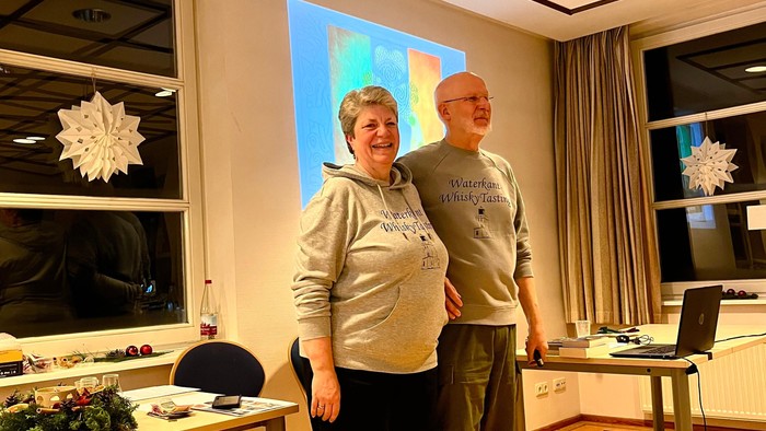 Zwei Personen stehen zusammen drinnen in der Nähe eines dekorierten Fensters und einer Präsentationsleinwand während einer Veranstaltung.