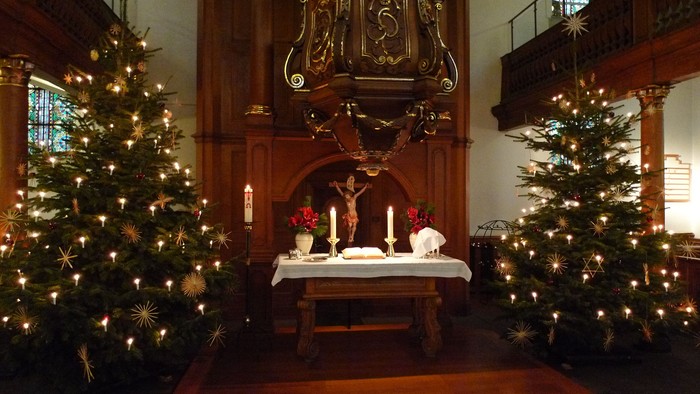 Zwei Weihnachtsbäume flankieren einen Altar mit Kerzen und Weihnachtsdekoration in einer Kirche.