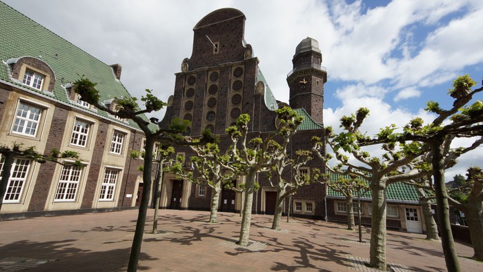 Das Bild zeigt einen großen Uhrturm mit Zifferblättern und umgebende Gebäude mit grünen Dächern und Bäumen.
