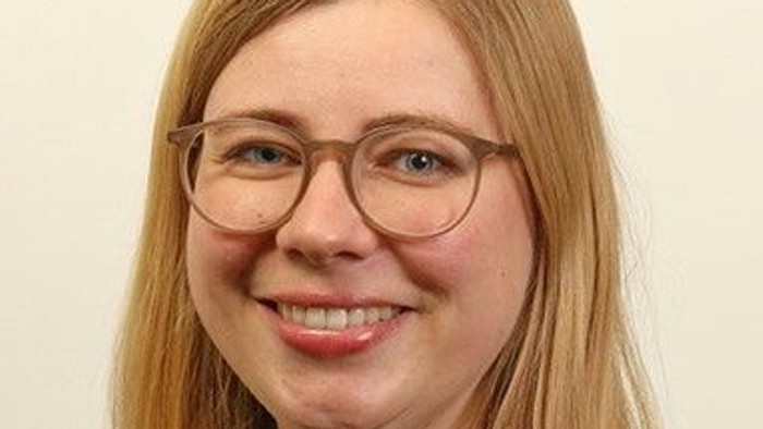 Lächelnde Frau mit blonden Haaren, die eine runde Brille trägt, vor einem schlichten Hintergrund.