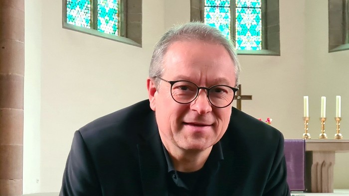 Mann in schwarzem Anzug und Brille, der in einer Kirche sitzt.