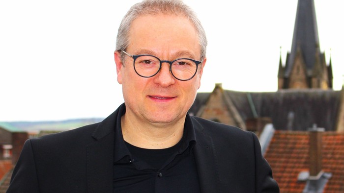 Ein Mann mit Brille und Sakko steht vor einer Kirche mit hohem Turmspitz.