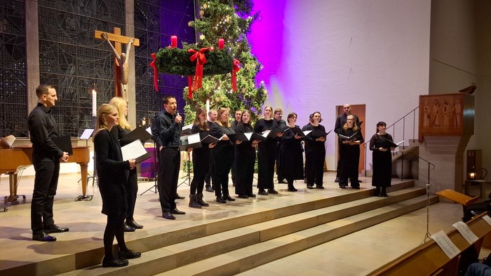 Ein gemischter Chor in schwarzer Chorkleidung in einem Altarraum, gesehen von vorne links. Über ihnen ein Adventskranz, im Hintergrund ein Weihnachtsbaum. 