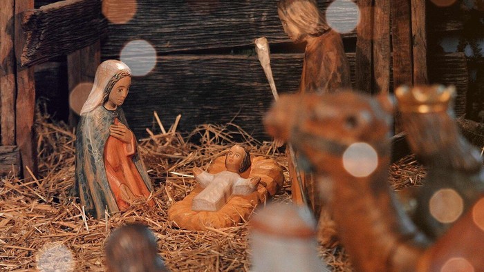 Eine klassische Weihnachtskrippe aus Holz, mit dem Fokus auf der heiligen Familie. Im Vordergrund verschwommen ein paar Köpfe und ein Kamel.