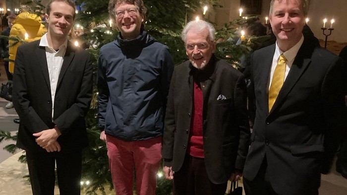 Vier Männer in formeller Kleidung posieren gemeinsam neben einem festlich geschmückten Weihnachtsbaum im Innenraum.