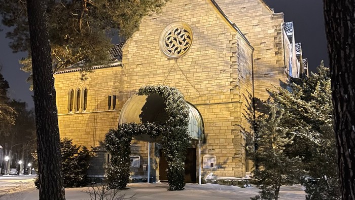 Eleganter Steinbau, nachts beleuchtet, mit einem kreisförmigen Emblem und einem gewölbten Eingang, umgeben von Bäumen.