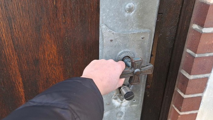 Eine Hand, die einen schweren Metalltürriegel an einer alten Holztür neben einer Backsteinmauer dreht.