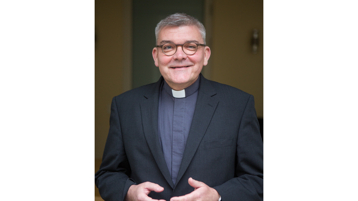 Ein Mann in geistlicher Kleidung mit Brille, der in der Nähe einer Tür steht und lächelt.