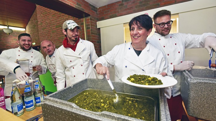 Köche, die gemeinsam in einer Küche eine große Portion grünen Gemüseeintopf zubereiten und servieren.