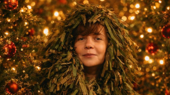 Eine Person mit einer festlichen Tannenzapfen-Perücke steht vor einem geschmückten Weihnachtsbaum mit funkelnden Lichtern.