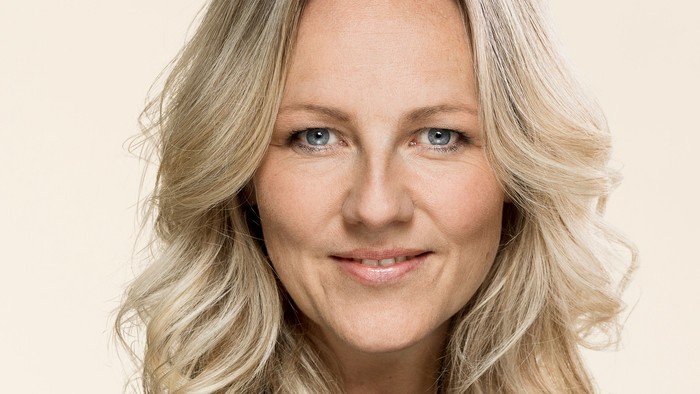 Professionelt portræt af en kvinde med blondt hår, der smiler mod en ensfarvet baggrund.