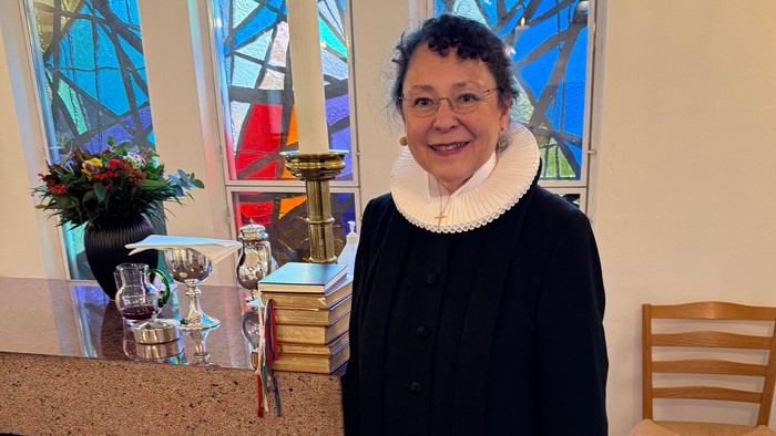 En person i religiøst tøj står ved et kirkealter med farvede glasvinduer bag sig.