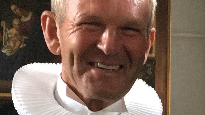 En smilende mand iført en sort kjole med en hvid krøllet krave, muligvis gejstligt tøj.