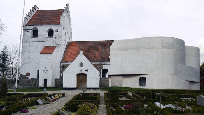 Historisk kirke med traditionel arkitektur ved siden af en moderne, buet betonbygning i en anlagt kirkegård.