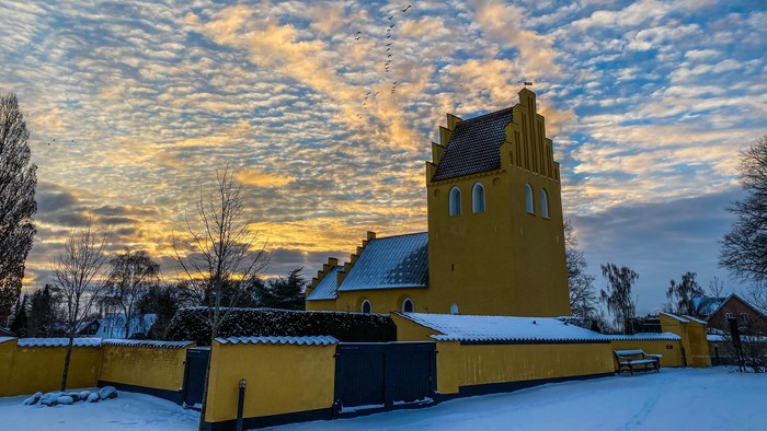 Gul kirke med tårn og spiret under snødekt himmel med solopgang