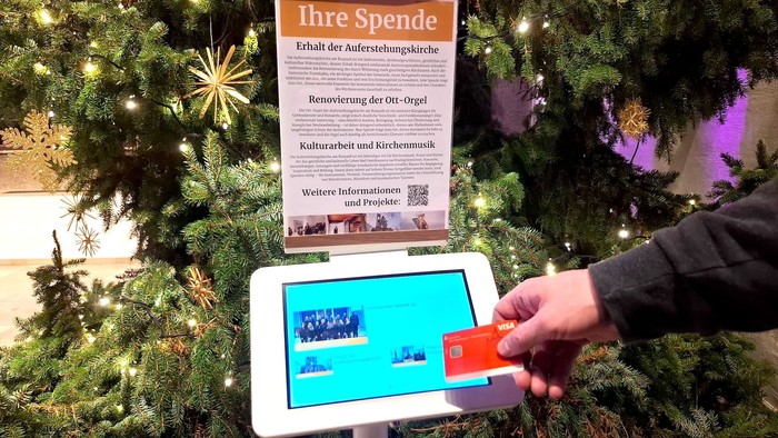 Eine Sparkassenkarte wird vor eine digitale Spendensäule gehalten. Über der Säule ein Blatt mit verschiedenen Spendenzwecken. Dahinter ein Weihnachtsbaum in Teilen zu erkennen.