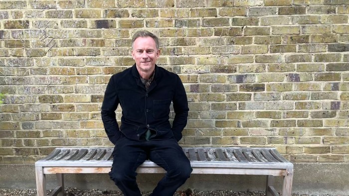 Mand i sort klædning sidder på bænk foran murstenge væg