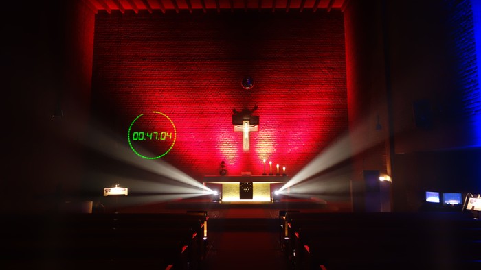 Auf dem Bild ist ein Altar mit darüber hängendem Kreuz zu sehen. Die Rückwand des Raumes leuchtet rot. Vom Altar selbst gehen weiße Lichtstrahlen aus, die in der Länge breiter werden. Ansonsten ist Kirchraum, in dem auch Bänke ein Reihen stehen, dunkel.