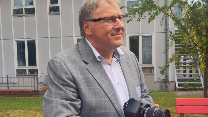 Ein Mann lächelt in die Kamera und hält in seinen Händen auch eine Kamera. 