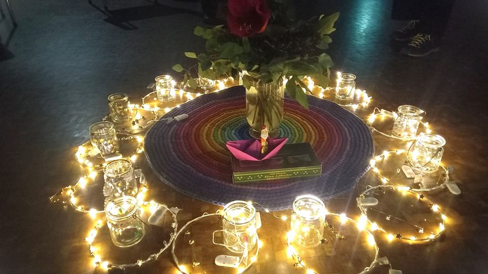 Elegante Tischdekoration mit Regenbogen-Häkelunterlage, Kerzen, Lichtern und einem Blumenarrangement als Mittelstück.