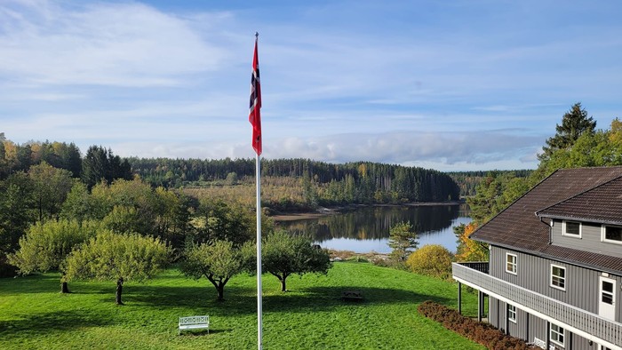 Malerisches Haus am See mit Fahnenmast, üppigem Grün und bewaldeten Hügeln im Hintergrund.