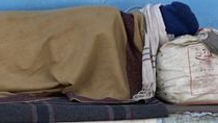 Eine Person schläft in einem minimalistischen Raum auf einem einfachen Stockbett mit einer Decke und einem Kissen.