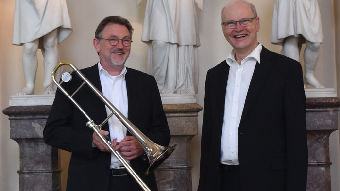 Zwei Männer posieren mit einer Posaune vor klassischen Statuen, beide formal in Anzügen und Krawatten gekleidet.