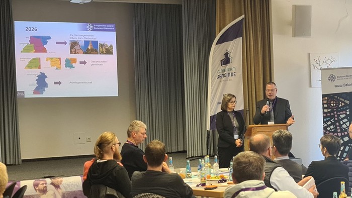 Ein Konferenzraum mit sitzenden Personen, ein Mann, der an einem Rednerpult spricht, und dahinter ein Präsentationsbildschirm.