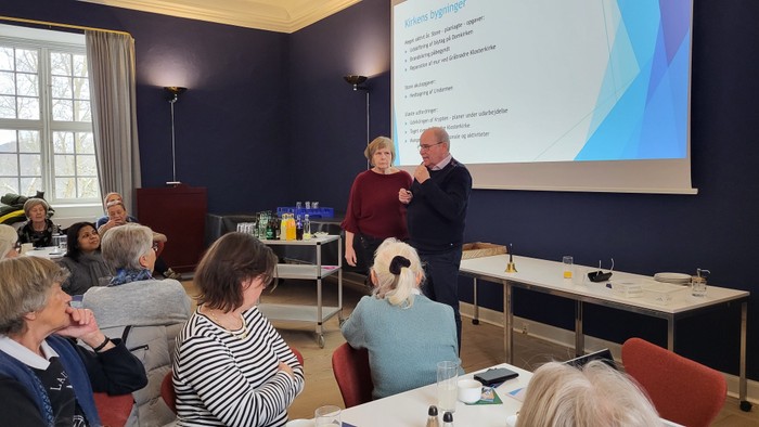 En gruppe mennesker deltager i en præsentation eller workshop med to oplægsholdere, der forklarer et emne på en skærm.