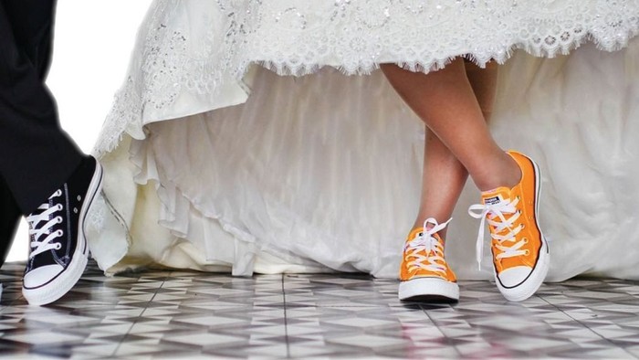 Ein verspieltes Paar steht auf einem gefliesten Boden, wobei eine Person ein Hochzeitskleid und orange Sneaker trägt, die andere schwarze Schuhe.