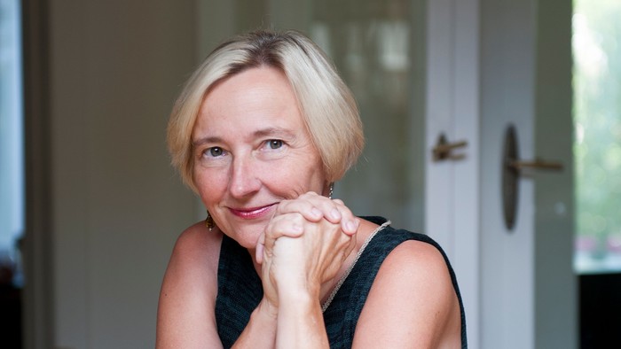 Eine Frau mit blonden Haaren sitzt nachdenklich an einem Holztisch in einer hellen Innenraumeinrichtung.