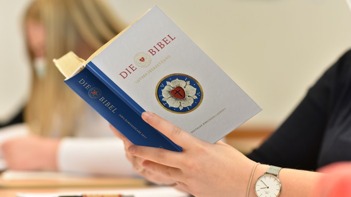 Eine Person hält ein blaues Buch mit dem Titel *„Die Bibel“* und einem floralen Design auf dem Umschlag.