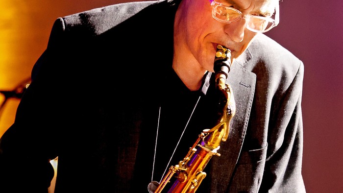 En mand med briller spiller saxofon på scenen med fokuseret intensitet.