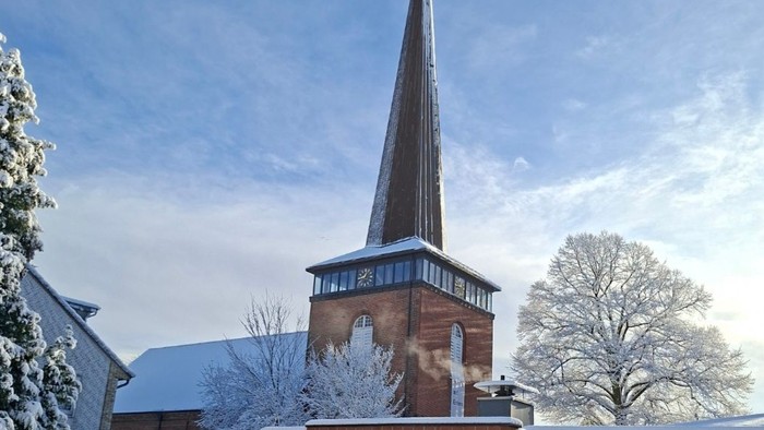 Eine schneebedeckte Kirchturmspitze ragt über eine winterliche Landschaft mit vereisten Hecken und kahlen Bäumen.