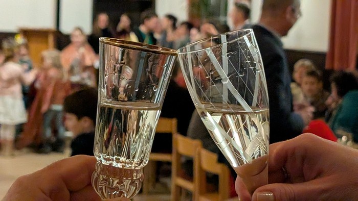 Zwei Hände, die Gläser mit klarem Getränk in einer festlichen Runde anstoßen, mit verschwommener Menschenmenge im Hintergrund.