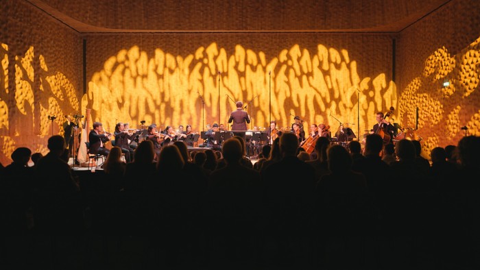 Ein Orchester spielt auf einer Bühne vor einem beleuchteten Hintergrund.