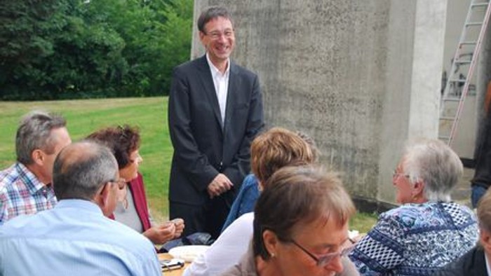 Eine Gruppe von Menschen im Freien, die sich um Tische versammelt hat, wobei ein Mann steht und zu ihnen spricht.