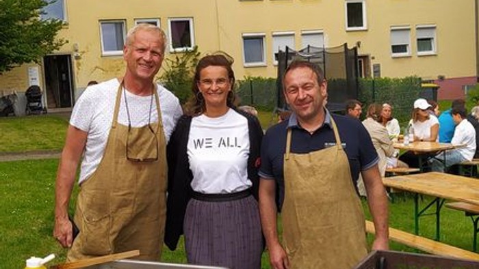 Drei Personen in Schürzen posieren gemeinsam bei einer Grillveranstaltung im Freien mit einem gelben Wohngebäude im Hintergrund.
