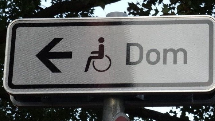 Straßenschild vor Baum: Pfeil, Pictogramm eines rollstuhlfahrers mit Rollstuhl, Dom.