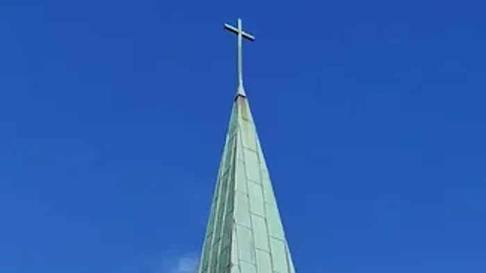 Kirche mit hohem grünem Turmspitz und Kreuz vor blauem Himmel.