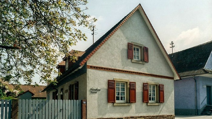 Das Bild zeigt ein charmantes, kleines Haus mit rotem Dach und weißen Fensterläden an einer Kopfsteinpflasterstraße.