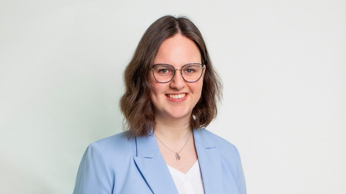 Professionelle Frau in einem hellblauen Blazer, mit Brille und einem warmen Lächeln vor einem schlichten Hintergrund.