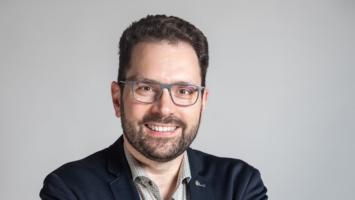 Ein Mann mit Brille und dunklem Jackett lächelt selbstbewusst.