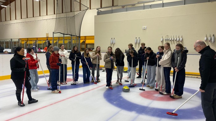 Gruppe, der øver curling indendørs på en isbane med spillere, der holder koste og sten.