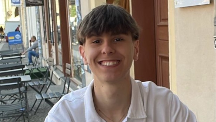 Eine Person, die vor einem Café mit Außenbestuhlung lächelt, während im Hintergrund Menschen zu sehen sind.